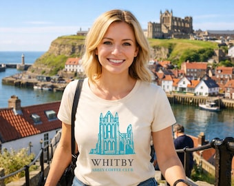 May include: A woman wearing a cream-colored t-shirt with a teal graphic of a building and the text "WHITBY ABBEY COFFEE CLUB." The background features a coastal town with a harbor, lighthouse, and a large building on a hill.