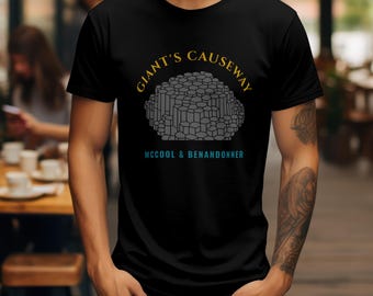 May include: Black t-shirt featuring a graphic of the Giant's Causeway, with text that reads "GIANT'S CAUSEWAY" in gold and "MCCOOL & BENANDONNER" in teal. The graphic depicts a cluster of gray hexagonal columns.