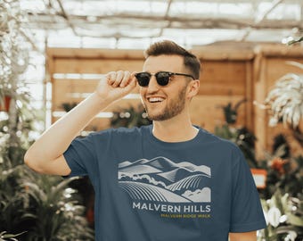 May include: Navy blue t-shirt with a white graphic of rolling hills and fields. The text "MALVERN HILLS" is above "MALVERN RIDGE WALK" in a yellow font. The shirt is being worn by a person wearing sunglasses.
