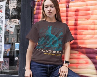 May include: A brown t-shirt featuring a teal illustration of a hare with the text "CAIRNGORMS MOUNTAIN HARE". The design includes mountain and plant elements. The shirt is made of a soft material.