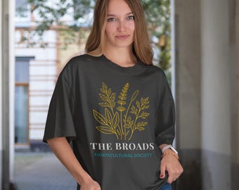 May include: A dark gray t-shirt featuring a gold botanical illustration and the text "THE BROADS HORTICULTURAL SOCIETY" in white and teal. The shirt has short sleeves and a relaxed fit.