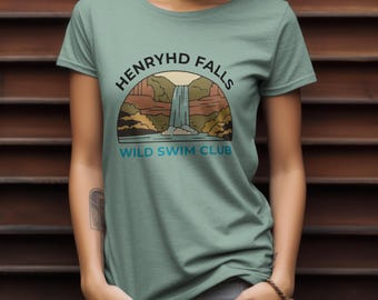 May include: A light green t-shirt featuring a graphic of a waterfall scene with the text "HENRYHD FALLS WILD SWIM CLUB" in a semi-circle above and below the image. The design includes a waterfall, mountains, and trees.