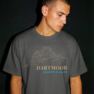 Könnte beinhalten: Graues T-Shirt mit einer beigen Strichzeichnung einer Felsformation. Der Text "DARTMOOR" steht über "GRANITE & GORSE" in Blaugrün. Das T-Shirt wird von einer Person getragen.