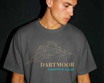 May include: A gray t-shirt featuring a tan line drawing of a rock formation. The text "DARTMOOR" is above "GRANITE & GORSE" in a blue-green color. The shirt is being worn by a person.