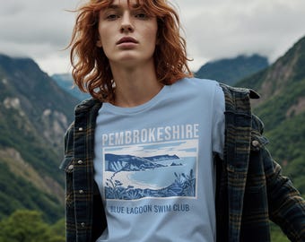 May include: A light blue t-shirt with the text "PEMBROKESHIRE" and "BLUE LAGOON SWIM CLUB" printed on the front. The graphic depicts a coastal scene in shades of blue and white. The person is wearing a plaid jacket.