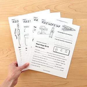 May include: A stack of white paper worksheets with black text and illustrations. The worksheets are titled "The Life of Elijah" and include fill-in-the-blank exercises and drawing prompts. The worksheets are held by a hand.