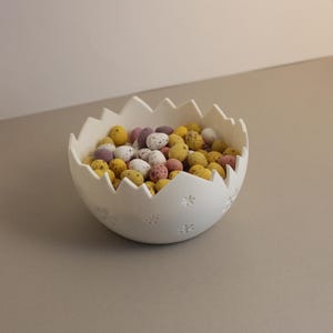 以下が含まれることがあります： ひびの入った卵の形をした白いセラミックボウル。カラフルな斑点模様のキャンディーが詰まっている。ボウルはギザギザの縁で、小さな浮き彫りの花の形で飾られている。キャンディーは黄色、白、ピンク、紫。