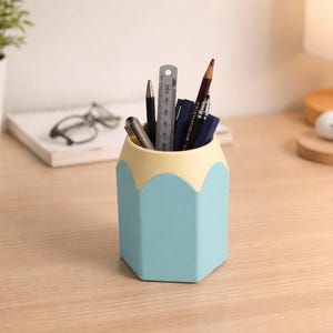 May include: A blue and cream pencil-shaped desk tidy containing pens, pencils, and a ruler. The organiser sits on a wooden desk, with a plant, glasses, and a lamp in the background. A keyboard is also visible.
