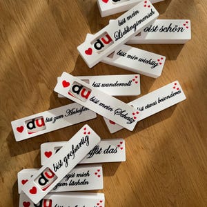 以下が含まれることがあります： 赤と黒の「du」の文字と赤いハートが付いた白い長方形の木製ブロック。各ブロックには、「bist perfekt」や「bist mein Schatz」など、黒いスクリプトで異なるドイツ語のフレーズが書かれています。ブロックは木製の表面に散らばっています。
