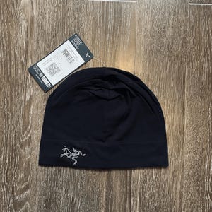 May include: A black beanie hat with a white logo on the front. The hat is made of a soft material and has a tag attached. The hat is laying on a wooden surface.