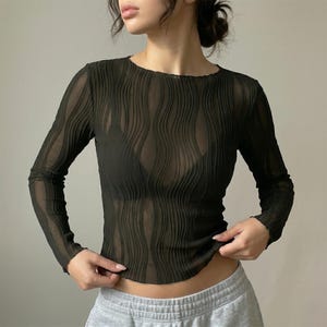 May include: Dark olive green long-sleeve top with a sheer, textured design. The top features a round neckline and a wavy, vertical pattern. The fabric appears lightweight and semi-transparent, suitable for layering or warmer weather.