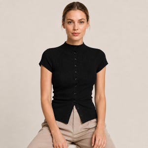 Puede incluir: Una prenda superior negra de manga corta con cuello alto y una hilera vertical de pequeños botones en la parte delantera. La prenda está confeccionada en tejido de punto acanalado que ofrece una silueta ajustada, terminando en la cintura.