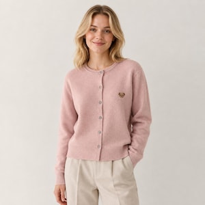 May include: A light pink long-sleeve cardigan with a round neckline and a button-down front. The garment features a small, embroidered brown bear head on the left chest area and ribbed cuffs and hem, paired with light beige trousers.