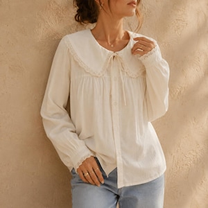 Pode incluir: Blusa creme com gola Peter Pan e mangas compridas. A gola e os punhos são adornados com renda delicada. A blusa tem uma frente abotoada e um detalhe de laço no decote. O tecido parece leve e ligeiramente texturizado.