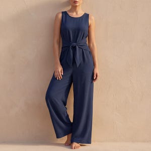 May include: A sleeveless, navy blue jumpsuit with a tie-waist detail. The wide-leg design and solid color create a simple, elegant look. The jumpsuit is made of a soft, flowing fabric, perfect for warm weather or casual occasions.