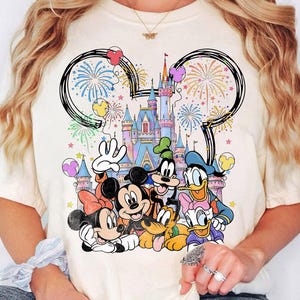 May include: Cream-colored t-shirt featuring a colorful graphic of Disney characters, including Mickey and Minnie Mouse, Goofy, Donald Duck, and Pluto, in front of a castle. The design includes fireworks and a Mickey Mouse ear outline.