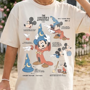 May include: Cream-colored t-shirt featuring a graphic of Mickey Mouse as the Sorcerer's Apprentice from Fantasia. The design includes illustrations and text detailing the character's features and the film's production. The shirt has a relaxed fit.