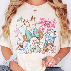 K&ouml;nnte beinhalten: Cremefarbenes T-Shirt mit farbenfrohem Grafikdesign. Das Design umfasst eine Schleife, Mickey Mouse Ohren, Popcorn, einen High Heel und den Text "Literally just a girl". Die Grafik ist in Rosa-, Blau- und Braunt&ouml;nen gehalten.