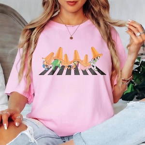 May include: A light pink t-shirt featuring a cartoon graphic of characters crossing a black and white striped crosswalk, each wearing an orange traffic cone. The characters include a green dinosaur, a pig, and Buzz Lightyear.