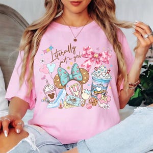 K&ouml;nnte beinhalten: Hellrosa T-Shirt mit einem farbenfrohen Grafikdesign mit Mickey Mouse Ohren, Schleifen und Lebensmitteln. Der Text "Literally just a girl" ist auf der Vorderseite aufgedruckt. Das T-Shirt hat kurze &Auml;rmel.