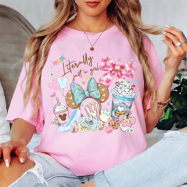 K&ouml;nnte beinhalten: Hellrosa T-Shirt mit einem farbenfrohen Grafikdesign mit Mickey Mouse Ohren, Schleifen und Lebensmitteln. Der Text "Literally just a girl" ist auf der Vorderseite aufgedruckt. Das T-Shirt hat kurze &Auml;rmel.
