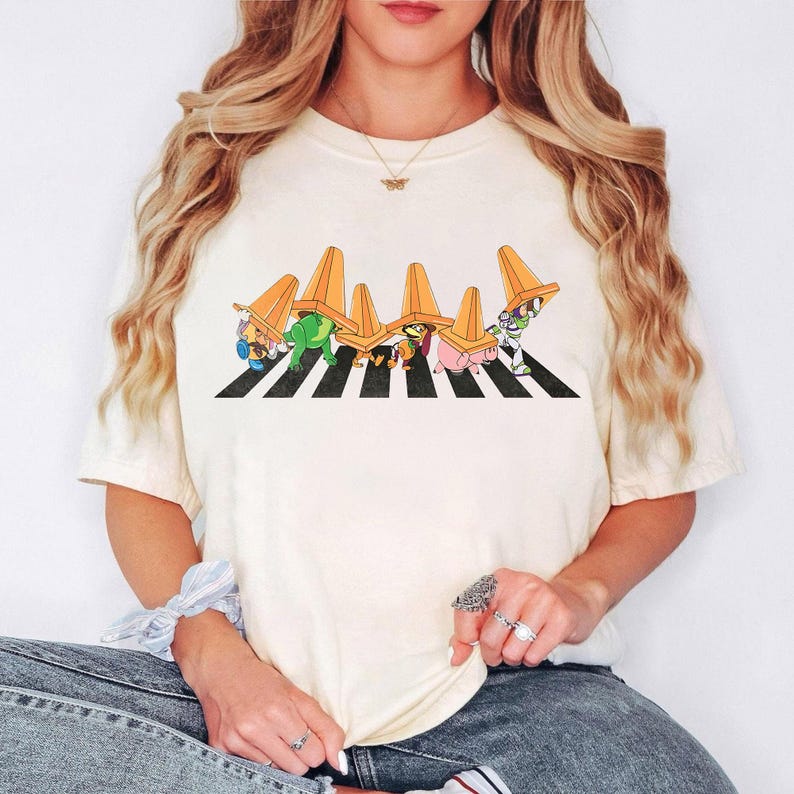 May include: Cream-colored t-shirt featuring a cartoon illustration of various characters wearing orange traffic cones and crossing a black and white crosswalk. The design is centered on the shirt.