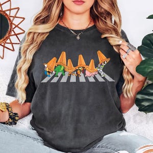 May include: Dark grey t-shirt featuring a cartoon parody of the Abbey Road album cover. The design shows characters crossing a crosswalk, each wearing an orange traffic cone. The characters include a turtle, a dinosaur, a dog, a pig, and Buzz Lightyear.