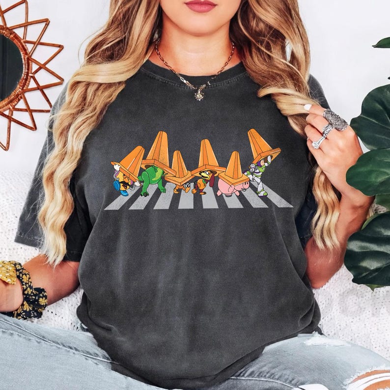 May include: Dark grey t-shirt featuring a cartoon parody of the Abbey Road album cover. The design shows characters crossing a crosswalk, each wearing an orange traffic cone. The characters include a turtle, a dinosaur, a dog, a pig, and Buzz Lightyear.