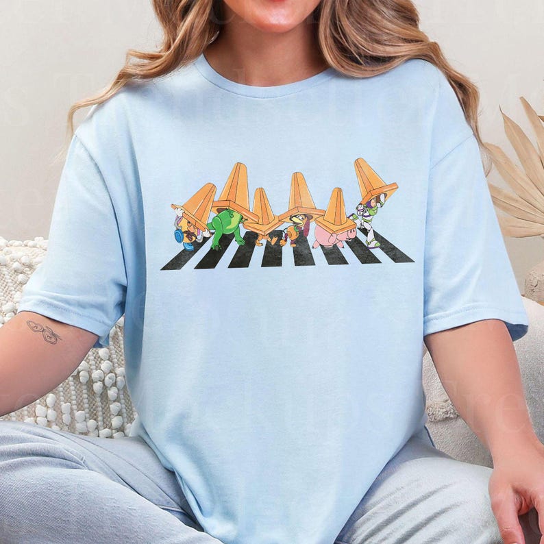 May include: Light blue t-shirt featuring a cartoon illustration of characters crossing a crosswalk, each wearing an orange traffic cone. The design is a parody of the iconic Abbey Road album cover.