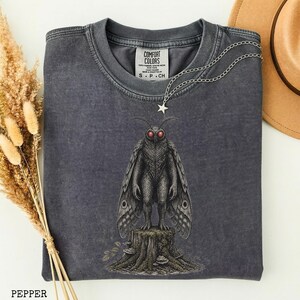 May include: A charcoal gray crew neck t-shirt featuring a graphic of a moth-like creature with red eyes standing on a tree stump. The shirt has a Comfort Colors label at the inner neckline and a silver-toned chain necklace with a star charm.