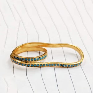 Puede incluir: Un anillo doble de tono dorado con una forma ovalada alargada y continua. La banda está incrustada con una serie de pequeñas piedras redondas de color turquesa brillante que siguen todo el perímetro del marco de metal.