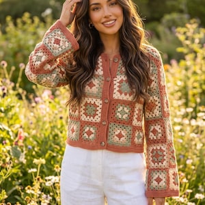 May include: A long-sleeved crochet cardigan featuring a patchwork of granny squares in shades of terracotta, sage green, and cream. The garment has a round neckline, a button-down front with five brown buttons, and a cropped, straight-cut hemline.