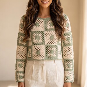 May include: A long-sleeved sweater constructed from alternating sage green and cream-colored crochet granny squares. The garment features a round neckline and a cropped hemline, paired with high-waisted, light beige trousers with front pleats.