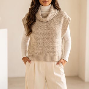 May include: A beige sleeveless crochet sweater vest featuring a wide cowl neckline and a ribbed hem. The garment is layered over a white long-sleeved turtleneck shirt and paired with cream-colored wide-leg trousers with front pleats.