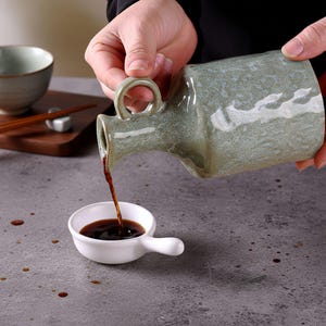 May include: A green and grey glazed ceramic sake bottle pours a dark liquid into a small white bowl with a handle. A bowl and chopsticks are visible on a wooden tray in the background. The scene suggests a Japanese dining setting.