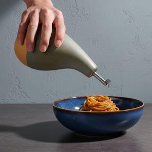 May include: A ceramic olive oil dispenser with a stainless steel spout pouring oil over a bowl of pasta. The dispenser is two-toned, with a green body and a tan base. The bowl is blue with a brown rim.