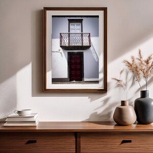 May include: A framed wall art print depicting a white building facade with a dark red door and a balcony with a metal railing. The print is displayed above a wooden sideboard featuring a stack of books, a white cup, and two vases with dried grass.