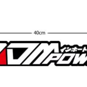 Puede incluir: Una calcomanía de vinilo negra, blanca y roja con el texto JDM POWER y caracteres japoneses sobre la palabra POWER. La calcomanía mide 40 cm de ancho y 6,5 cm de alto, según lo indicado por las líneas de dimensión visibles.