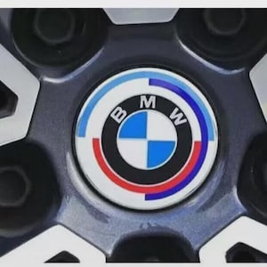 Può includere: Primo piano di un coprimozzo BMW. Il tappo presenta il logo BMW, un emblema circolare con anelli concentrici blu, bianchi e neri e le lettere "B", "M" e "W". La ruota ha una finitura grigio scuro.