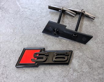 Könnte beinhalten: Ein schwarz-rotes S6-Emblem mit glänzender Oberfläche, bei dem der Buchstabe S und die Zahl 6 in Schwarz vor einem roten rechteckigen Hintergrund stehen. Das Emblem enthält eine schwarze Halterung mit zwei Gewindestangen und Metall-Kippdübeln.