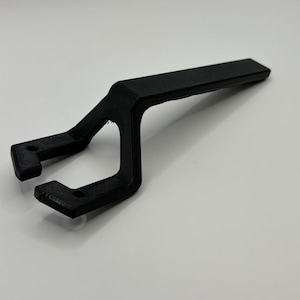 May include: A matte black, 3D-printed plastic tool featuring a long, straight handle that transitions into a Y-shaped head. The two prongs of the head each contain a small, circular hole, designed for mechanical alignment or adjustment tasks.