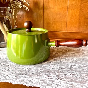 May include: A green enamel saucepan with a wooden handle and lid. The handle and lid knob are a dark brown color. The saucepan is sitting on a white lace tablecloth.