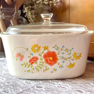 May include: White casserole dish with a clear glass lid. The dish is decorated with a floral pattern featuring orange, yellow, pink, and blue flowers. The dish has two handles and is likely used for cooking and serving.
