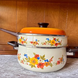 Puede incluir: Juego de olla y vaporera de esmalte vintage de color crema con diseños florales en rojo, amarillo, azul y naranja. La tapa es naranja con un asa marrón oscuro. El juego tiene asas marrones y está sobre un mantel de encaje blanco.