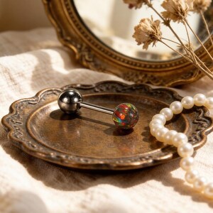 Könnte beinhalten: Ein Zungenpiercing im Barbellen-Stil mit einem glänzenden silberfarbenen Metallschaft und zwei Kugelenden. Ein Ende ist eine massive silberne Kugel, und das andere ist eine farbenfrohe, irisierende Opal-Kugel. Der Schmuck liegt auf einer dekorativen Metallplatte im antiken Stil.