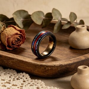 Könnte beinhalten: Ein schwarzer Ring mit einem farbenfrohen Inlay aus roten, blauen und lila Steinen. Der Ring liegt auf einem Holztablett mit einer getrockneten Rose und kleinen dekorativen Vasen. Das Innenband des Rings ist goldfarben.