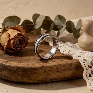 Könnte beinhalten: Ein silberner Ring mit schwarzem und weißem Inlay. Der Ring wird auf einer Holzoberfläche mit einer getrockneten Rose und Grünzeug im Hintergrund präsentiert. Das Design des Rings weist ein zentrales schwarzes Band auf, das von weißen Bändern flankiert wird.