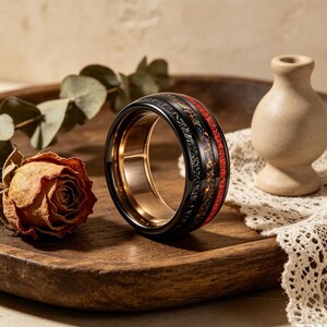 Könnte beinhalten: Ein schwarzer Ring mit goldener Innenseite und einem dekorativen Band mit roten und mehrfarbigen Akzenten. Der Ring wird auf einem Holztablett mit einer getrockneten Rose und einer kleinen Vase präsentiert.