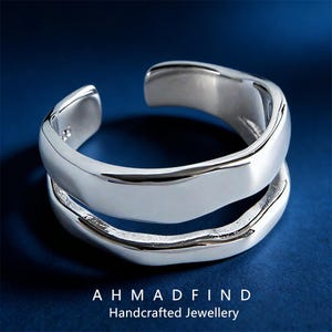 May include: A silver-colored ring with a unique, open design. The ring features two bands that curve and overlap, creating a modern and stylish look. The ring is displayed on a dark blue background. Text at the bottom reads "AHMADFIND Handcrafted Jewellery."