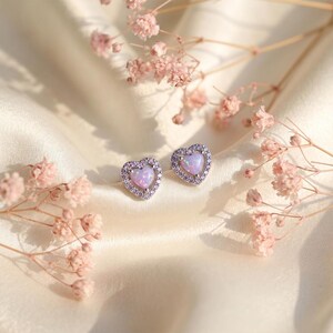 May include: A pair of heart-shaped stud earrings. Each earring features a light purple opal center stone surrounded by small, clear crystals. The earrings are set on a silver-colored metal and rest on a cream-colored fabric, with pink flowers in the background.